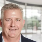 Tom Griga headshot on blurred background of corporate lobby with trees in the distance