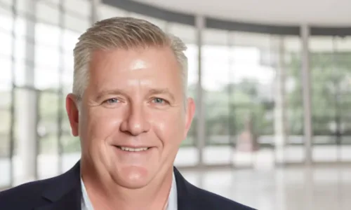 Tom Griga headshot on blurred background of corporate lobby with trees in the distance